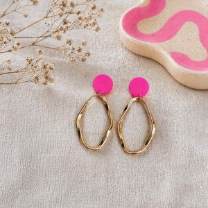 Könnte beinhalten: Ein Paar goldfarbene, unregelmäßig geformte Creolen mit leuchtend pinkfarbenen, kreisförmigen Nieten. Im Hintergrund befindet sich eine kleine, rosa-weiße Keramikschale mit einem wellenförmigen rosa Design. Die Ohrringe liegen auf einem neutralfarbenen Stoff.