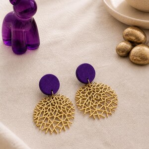 Könnte beinhalten: Ein Paar goldfarbene, offene Ohrringe mit kreisförmigem Design, jeweils befestigt an einem kleinen, runden, lila Ohrstecker. Eine lila Bärenfigur und goldfarbene dekorative Steine sind ebenfalls zu sehen.
