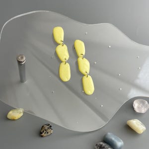 May include: A pair of yellow, oblong earrings with a glossy finish. The earrings are connected by small metal hoops, creating a dangling effect. The earrings are displayed on a clear, wavy surface with small decorative stones.