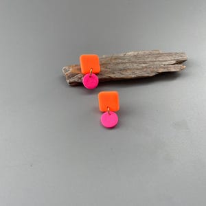Könnte beinhalten: Ein Paar Ohrringe mit neonorangen Quadraten und kleinen, hellrosa Kreisen darunter. Die Ohrringe sind auf einem Stück Treibholz vor grauem Hintergrund platziert. Die Ohrringe haben ein modernes, geometrisches Design.