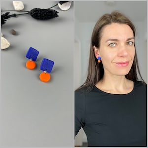 Könnte beinhalten: Ein Paar geometrische Ohrringe in Blau und Orange. Die Ohrringe sind aus Polymer Clay gefertigt und haben eine quadratische Form mit einem runden Anhänger.