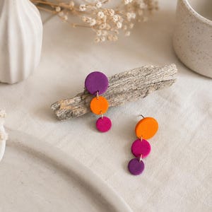 Könnte beinhalten: Ein Paar handgefertigte Ohrringe mit einem kaskadierenden Design aus farbigen Kreisen. Die Ohrringe zeigen Lila, Orange und Fuchsia, die an einem Stecker befestigt sind. Die Ohrringe werden auf einem Stück Treibholz präsentiert.