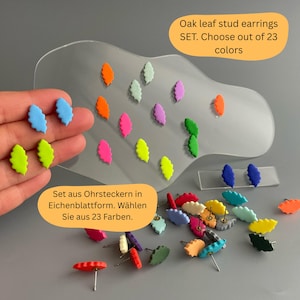 Peut inclure: Des boucles d'oreilles clous en forme de feuille de chêne, disponibles en plusieurs couleurs vives : bleu, vert, rose et orange. L'image présente plusieurs ensembles de boucles d'oreilles, certains disposés sur un présentoir transparent et d'autres éparpillés. Le texte indique : "Oak leaf stud earrings SET. Choose out of 23 colors."