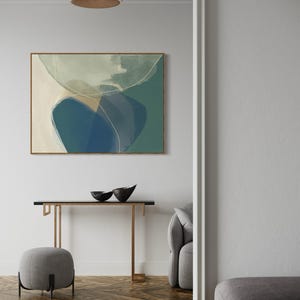 May include: A modern living room interior with a contemporary abstract painting featuring shades of blue, green, and white. The painting is framed in a light wood frame and hangs on a white wall. A console table with a black top and gold metal legs sits in front of the painting. Two black bowls are placed on the table. A gray ottoman sits in front of the table. A gray sofa is visible in the background.