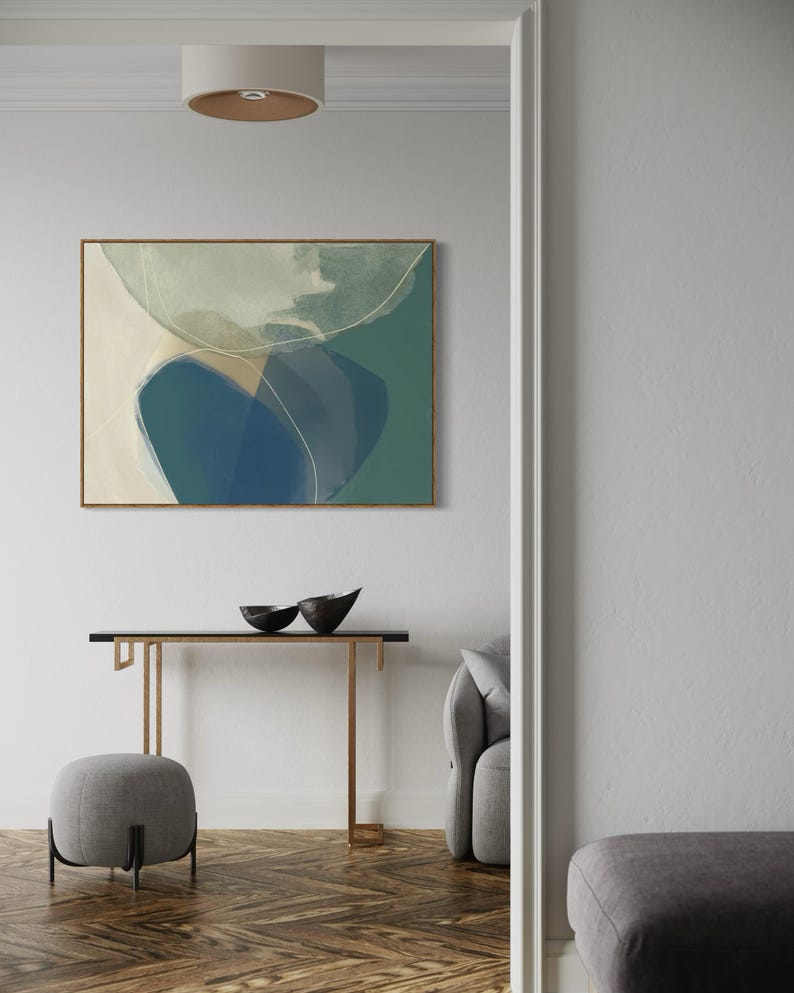 May include: A modern living room interior with a contemporary abstract painting featuring shades of blue, green, and white. The painting is framed in a light wood frame and hangs on a white wall. A console table with a black top and gold metal legs sits in front of the painting. Two black bowls are placed on the table. A gray ottoman sits in front of the table. A gray sofa is visible in the background.