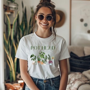 May include: White t-shirt with the text "POT HEAD" printed in green letters. The shirt features a graphic of a row of potted plants with different types of greenery.