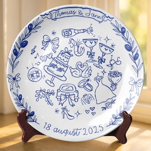 May include: White ceramic plate with blue hand-drawn illustrations of wedding-related icons, including a cake, champagne, and rings. The plate has a blue border with olive branches and the names "Thomas & Jane" and the date "18 august 2025".