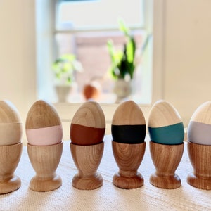 May include: Seven wooden eggs painted in different colors, each sitting in a wooden egg cup. The colors are natural wood, pink, brown, black, teal, and gray.