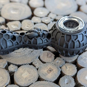 Puede incluir: Pipa para fumar de silicona negra con un patrón de panal y un diseño de abeja. La pipa tiene una cazoleta de vidrio transparente.