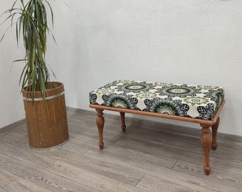 May include: A wooden bench with a patterned cushion featuring green and white floral designs. The bench has four curved legs and a light brown wooden frame. A tall potted plant is to the left.