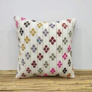 May include: White throw pillow with a colorful geometric pattern. The pattern features small, multi-colored flowers in a diamond shape. The pillow is sitting on a woven bamboo surface.