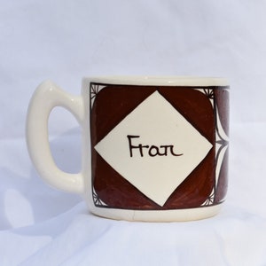 Peut inclure: Une tasse en céramique blanche avec un motif géométrique brun et le mot "Fran" écrit en noir à l'intérieur d'un losange.