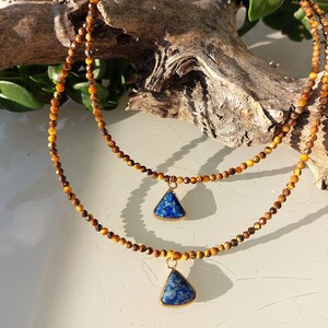 Puede incluir: Dos collares superpuestos con detalles dorados. Los collares están hechos de cuentas de ojo de tigre y presentan colgantes de lapislázuli azul.