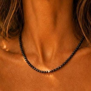 Può includere: Una collana composta da piccole perle sferiche nere opache, uniformi e infilate su un unico filo. Le perle sono costanti per dimensioni e forma, creando un design circolare semplice e continuo che riposa attorno alla zona del collo.