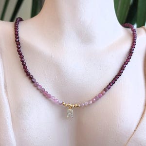 Peut inclure: Un collier délicat composé de petites perles facettées, de couleur violet foncé et rose. Le collier est orné d'un élément doré et d'un petit pendentif étincelant en forme de lettre "A". Le collier est présenté sur un mannequin.