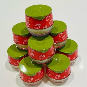May include: A stack of eight small, round containers with green lids and red bodies. The red bodies are decorated with white strawberry illustrations. The containers are stacked in a pyramid shape, with the lids facing up.