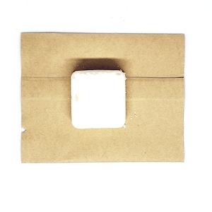 May include: A square, white soap bar sits on a brown paper background. The soap has a slightly textured surface. The paper is folded and creased. The background is white.