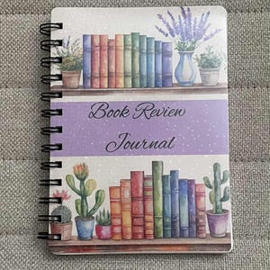 Puede incluir: Un cuaderno de espiral titulado Book Review Journal que presenta una ilustraci&oacute;n en acuarela de dos estantes de madera llenos de libros coloridos, cactus en macetas y un jarr&oacute;n con flores de lavanda sobre un fondo morado claro y blanco moteado.