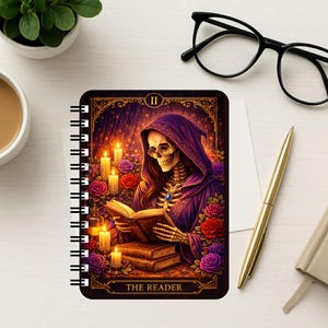Puede incluir: Un cuaderno de espiral con una ilustraci&oacute;n estilo tarot de un esqueleto con t&uacute;nica morada con capucha leyendo un libro. La imagen incluye velas encendidas, rosas rojas y moradas, y el texto THE READER en la parte inferior en letras doradas.