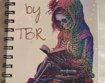 Puede incluir: Un cuaderno de espiral con acabado de estrellas holográficas que muestra un esqueleto con una capa rosa con capucha mientras lee un libro. El texto Death by TBR está impreso en marrón en el lado izquierdo sobre una pila de varios libros.