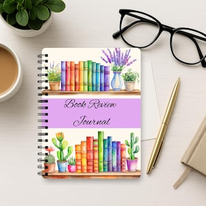 Puede incluir: Un diario de rese&ntilde;as de libros con encuadernaci&oacute;n en espiral que presenta una ilustraci&oacute;n en acuarela de dos estantes de madera llenos de libros coloridos, cactus en macetas y lavanda. El centro de la portada tiene una banda horizontal de color violeta claro con el texto Book Review Journal.