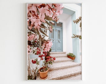 May include: A white building with a blue door and pink flowers growing on the side. The door is open and there are steps leading up to it. There are potted plants on the steps.