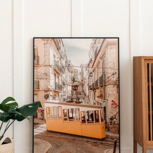 May include: A yellow tram car tilted on a cobblestone street in a city with tall buildings. The tram is facing the camera and is partially obscured by a building on the right side of the image.