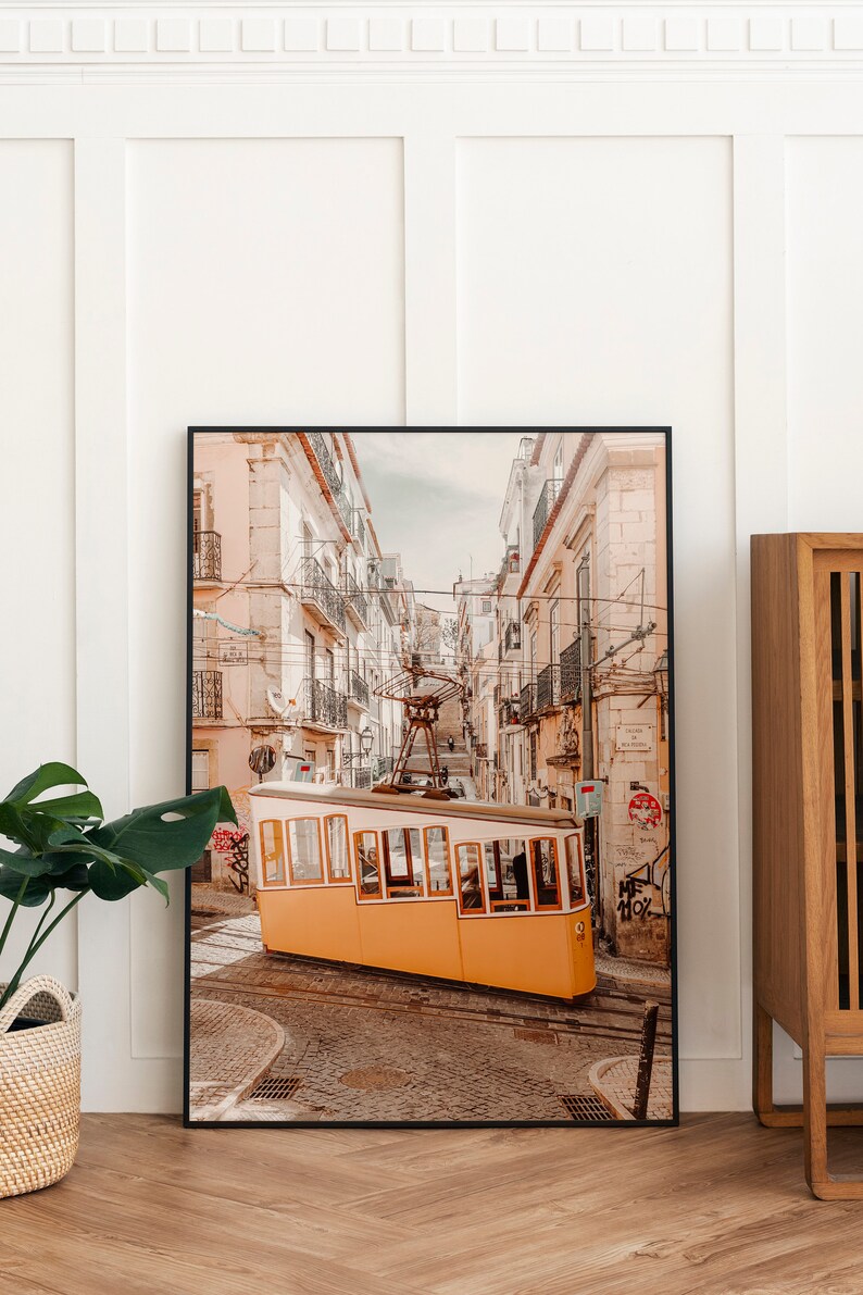 May include: A yellow tram car tilted on a cobblestone street in a city with tall buildings. The tram is facing the camera and is partially obscured by the buildings. The street is narrow and has a few people walking on it.