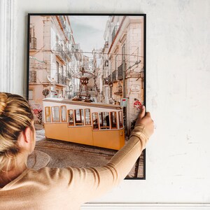 May include: A woman is hanging a framed photograph of a yellow tram in a narrow street in Lisbon, Portugal. The tram is going uphill and the street is lined with buildings.