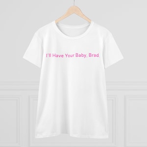 Puede incluir: Camiseta blanca con el texto "I'll Have Your Baby, Brad" en rosa. La camiseta está colgada en una percha de madera. La camiseta tiene cuello redondo y mangas cortas.