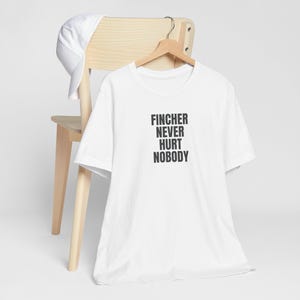 May include: White t-shirt with black text that reads "Fincher Never Hurt Nobody". The shirt is hanging on a wooden chair.
