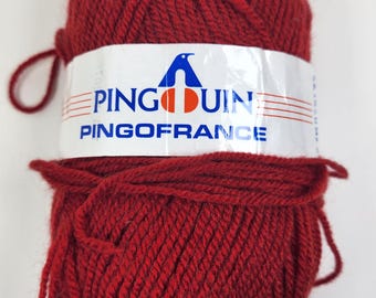 May include: A ball of deep red yarn with a white label wrapped around it. The label features a blue penguin logo and the text "PINGOUIN PINGOFRANCE". The yarn is tightly wound and ready for crafting.