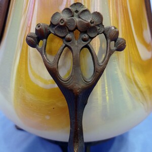 Puede incluir: Primer plano de un jarr&oacute;n de vidrio decorativo con un patr&oacute;n de remolino de blanco y amarillo. El jarr&oacute;n tiene un asa de bronce con un dise&ntilde;o floral.