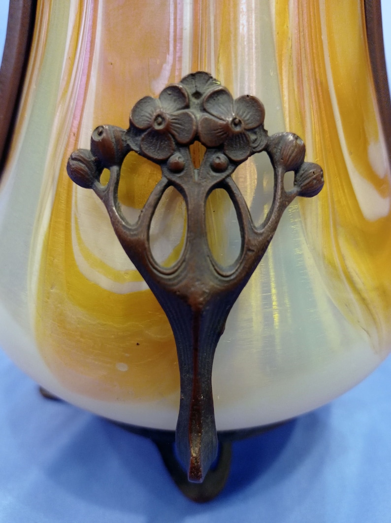 Puede incluir: Primer plano de un jarr&oacute;n de vidrio decorativo con un patr&oacute;n de remolino de blanco y amarillo. El jarr&oacute;n tiene un asa de bronce con un dise&ntilde;o floral.