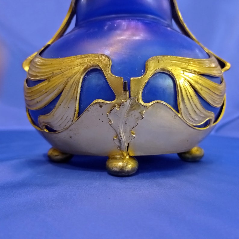 May include: A blue glass vase with gold accents and a decorative floral design. The vase has three feet and is made in the Art Nouveau style.