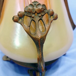 Puede incluir: Un jarr&oacute;n de vidrio vintage con un acabado iridiscente en espiral y una base de metal bronce. La base presenta un dise&ntilde;o floral decorativo con tres patas.