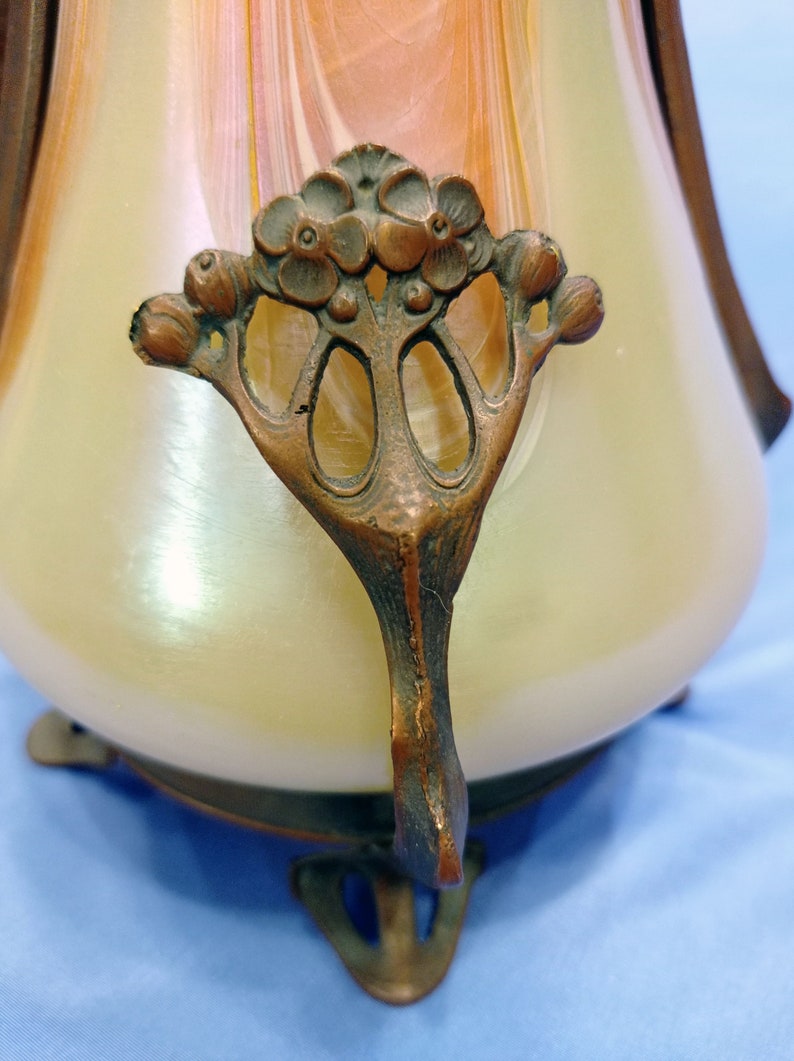 Puede incluir: Un jarr&oacute;n de vidrio vintage con un acabado iridiscente en espiral y una base de metal bronce. La base presenta un dise&ntilde;o floral decorativo con tres patas.