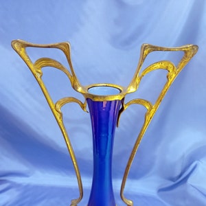 May include: A blue iridescent glass vase with a gold metal stand. The stand has a decorative design with intricate curves and swirls.