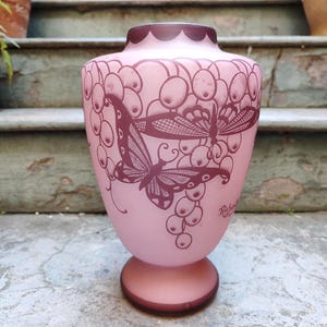Puede incluir: Un jarrón de cristal rosa con base redonda y cuerpo ancho. El jarrón presenta un borde festoneado morado oscuro y está decorado con mariposas grabadas y racimos de uvas. El jarrón mide aproximadamente 25 cm de alto.
