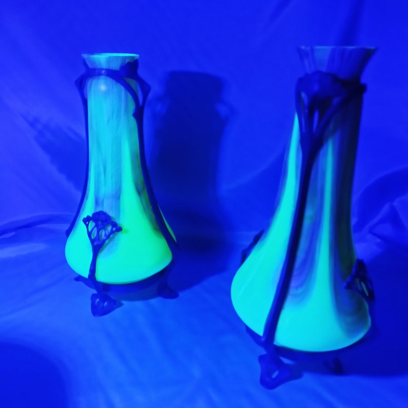 Puede incluir: Dos jarrones de vidrio verde con detalles en metal negro y dise&ntilde;os florales. Los jarrones est&aacute;n sobre un fondo azul y brillan bajo una luz negra.