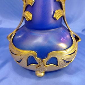 May include: A blue iridescent glass vase with a gold metal base. The base features an intricate design of stylized leaves and flowers.