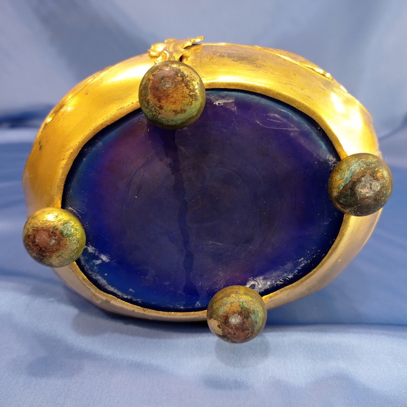May include: A gold-colored metal bowl with a blue glass bottom and four feet. The bowl has a decorative design around the rim.