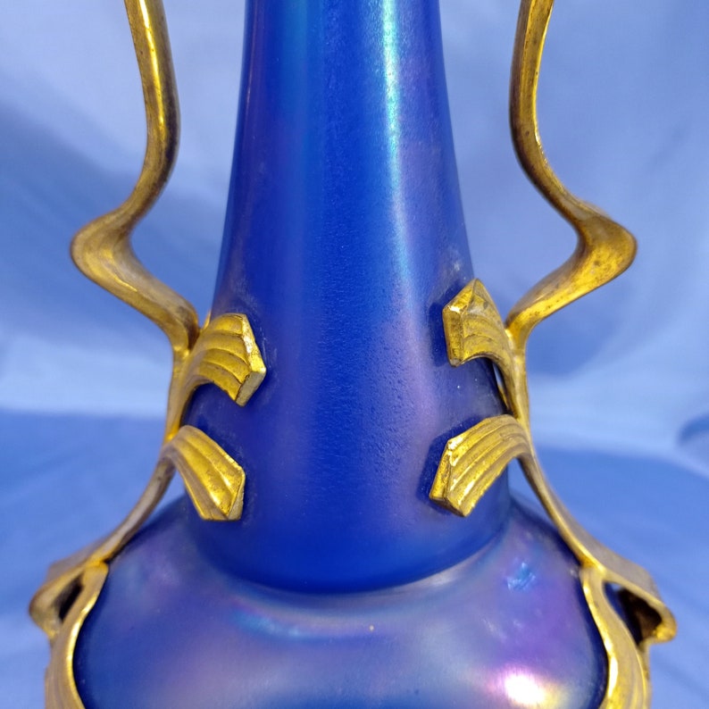 May include: A blue iridescent glass vase with gold metal accents. The vase has a rounded body and a long, thin neck. The gold accents are in the shape of leaves and vines.