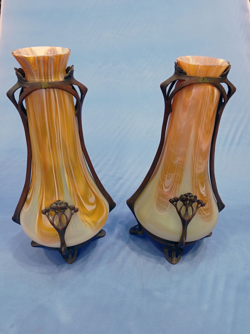 Puede incluir: Un par de jarrones antiguos de estilo art nouveau con un dise&ntilde;o de vidrio &aacute;mbar y blanco en espiral. Los jarrones tienen una base de metal bronce con un dise&ntilde;o floral.