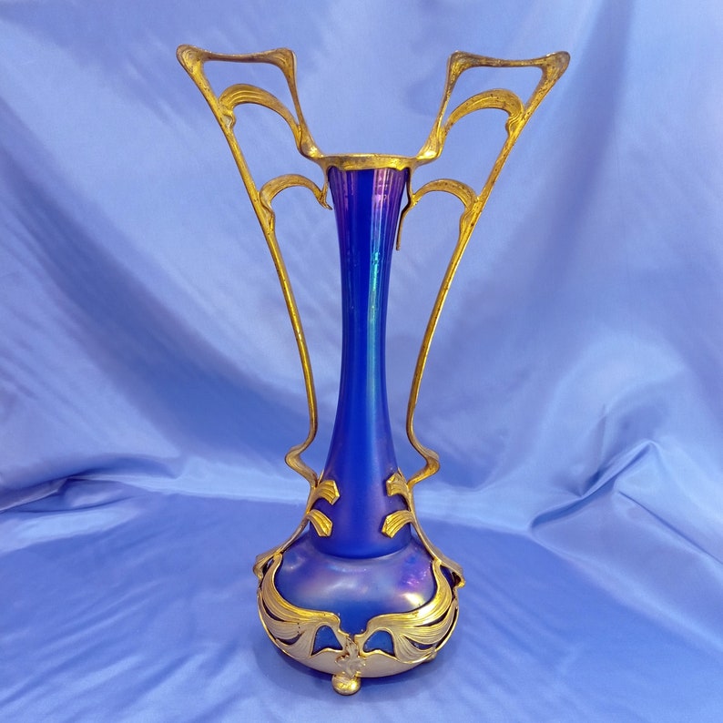 May include: A blue iridescent glass vase with a gold metal frame and ornate details. The vase has a tall, slender neck and a wide, rounded base.