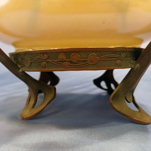 Puede incluir: Un macetero de cer&aacute;mica vintage con un esmalte amarillo p&aacute;lido y una base de bronce decorativa. La base presenta un dise&ntilde;o floral con hojas y flores estilizadas.