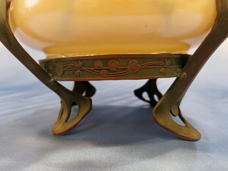 Puede incluir: Un macetero de cer&aacute;mica vintage con un esmalte amarillo p&aacute;lido y una base de bronce decorativa. La base presenta un dise&ntilde;o floral con hojas y flores estilizadas.