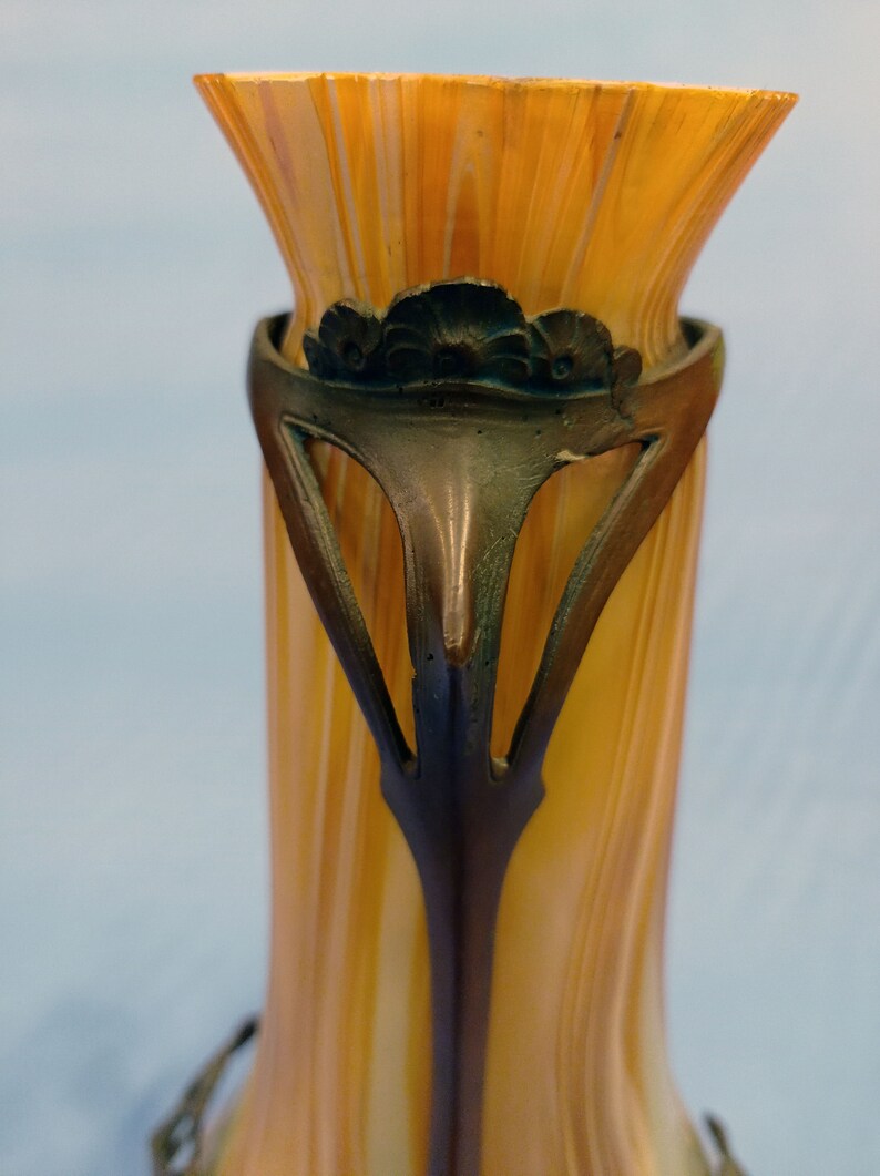 Puede incluir: Un jarr&oacute;n de vidrio alto de color &aacute;mbar con una base de metal bronce decorativa. La base presenta un dise&ntilde;o floral y recortes intrincados.
