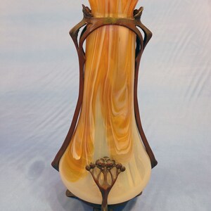 Puede incluir: Un jarr&oacute;n vintage de estilo art nouveau con un cuerpo de vidrio blanco y &aacute;mbar veteado y un marco de metal bronce. El jarr&oacute;n tiene una abertura acampanada y una base redondeada con tres patas.