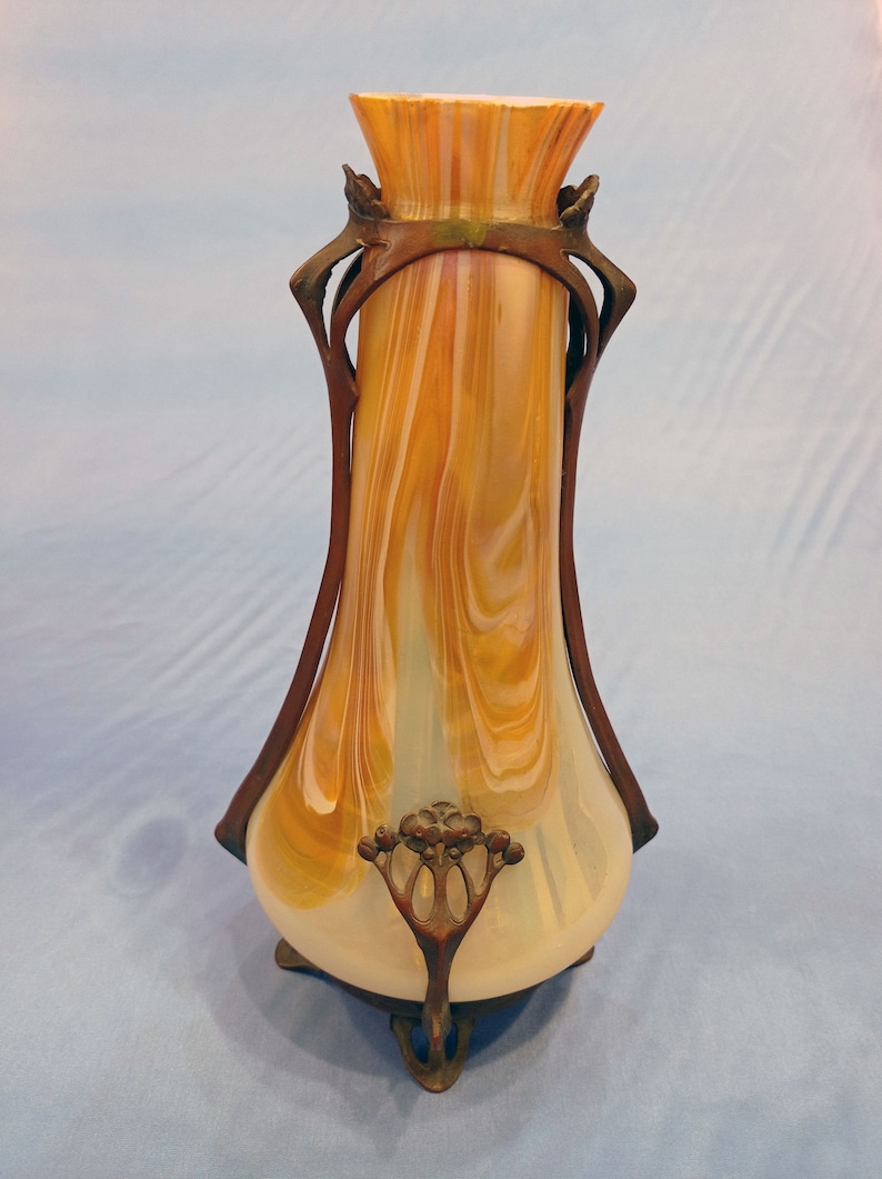 Puede incluir: Un jarr&oacute;n vintage de estilo art nouveau con un cuerpo de vidrio blanco y &aacute;mbar veteado y un marco de metal bronce. El jarr&oacute;n tiene una abertura acampanada y una base redondeada con tres patas.